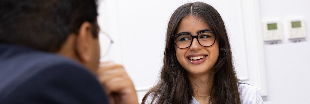 smiling female law student wearing business attire with glasses, looking at male law student wearing a suit and glasses.